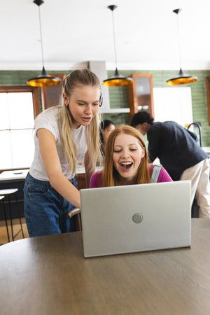 Teen girls laughing while watching something on laptop in modern kitchen. Friendship, technology,...