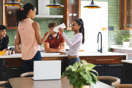 Teen girl receiving card from woman in kitchen, friends smiling in background. Friendship, celebration, happiness, gift, greeting, groupの写真素材