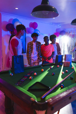 Teens celebrating around pool table with gift bags and colorful balloons. Celebration, party, enjoyment, friendship, leisure, funの写真素材