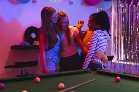 Teen girls laughing and hugging near pool table at vibrant party celebration. friendship, youth,...