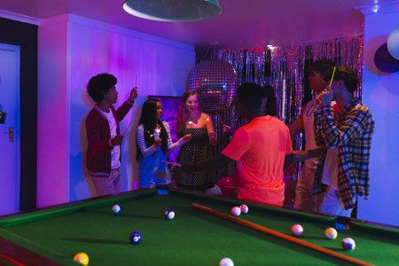 Teenagers celebrating around pool table with colorful lights and festive decorations. Party,...