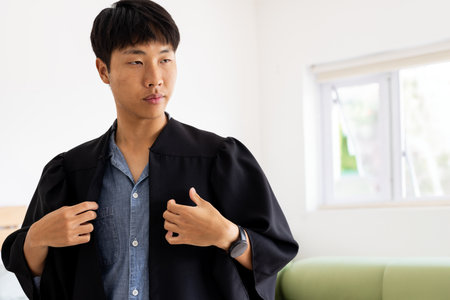 Young adult Asian man adjusting academic gown in white room with green sofa, copy space. Graduation, scholar, achievement, learning, preparation, minimalist, modernの写真素材