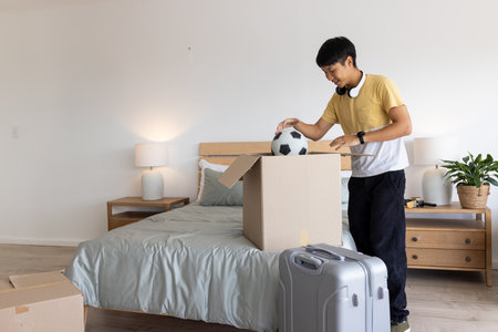 Asian man unpacking soccer ball from cardboard box in bedroom with rolling suitcase, copy space....