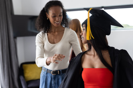 Proud mother helping daughter with graduation gown at home, celebrating achievement. ceremony, family, celebration, accomplishment, milestone, educationの写真素材