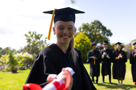 Graduate in cap and gown receiving diploma outdoors, smiling with friends applauding. Graduation,...