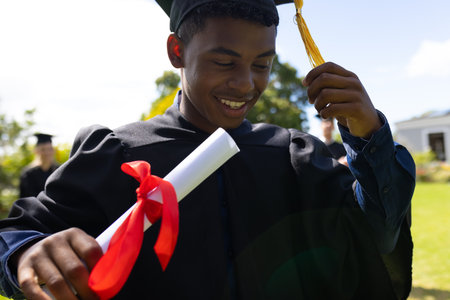 Teen graduate smiling outdoors, holding diploma with red ribbon, celebrating success. Graduation, celebration, achievement, education, happyの写真素材