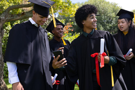 Graduating students taking selfies and holding diplomas, celebrating outdoors. Graduation, celebration, ceremony, joy, success, achievementの写真素材
