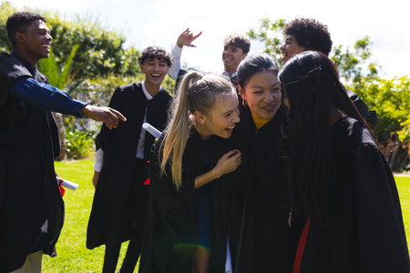 Teen graduates celebrating outdoors, laughing and embracing in graduation gowns. celebration, success, achievement, joy, friendship, educationの写真素材