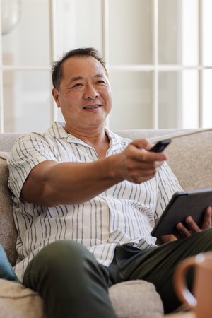 Asian man relaxing on sofa using remote control and holding tablet at home. Technology, leisure,...