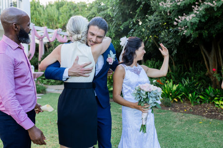 Groom hugging guest warmly while bride holding bouquet, smiling in garden. Wedding, celebration, joy, happiness, love, coupleの写真素材