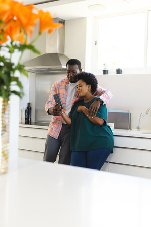 In kitchen, African American couple looking at smartphone, smiling together. Technology, lifestyle, cooking, happiness, connection, leisureの写真素材