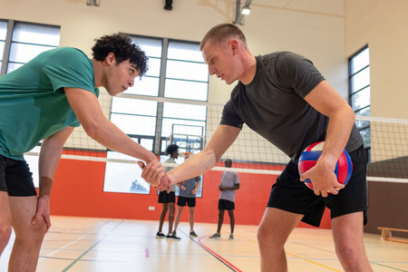 Male volleyball players shaking hands at net in school gym near bench, ball and court markings. Athleticism, teamwork, camaraderie, competition, focus, determination, vibrantの写真素材