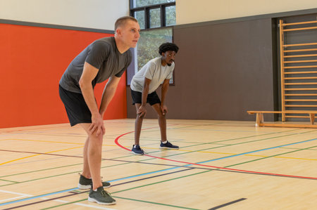 Diverse male teammates bending forward and resting on court with colored lines inside gymnasium....
