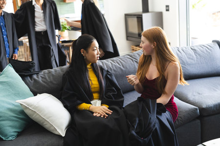 Teen friends in graduation gowns sitting on couch, talking and celebrating together. celebration, friendship, achievement, caps, classmates, joyの写真素材