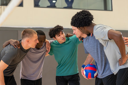 Diverse male teammates huddling together sharing pre-game strategy on gym court with volleyball. Athletes, teamwork, cohesion, fitness, sportsmanship, determination, camaraderieの写真素材