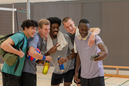 Diverse male friends posing for selfie on volleyball court with smartphone and volleyball. Athletic, camaraderie, fitness, teamwork, vibrant, active, recreationの写真素材