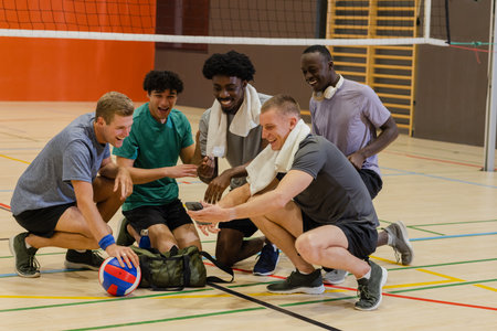 Diverse male volleyball players kneeling in gym under net laughing around smartphone and volleyball. Athletics, teamwork, camaraderie, fitness, recreation, vibrant, youthfulの写真素材