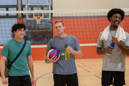 Diverse male friends standing on gym wood floor court holding volleyball water bottles and gym bag. Athletes, sports, teamwork, fitness, activewear, gymnasium, competitionの写真素材