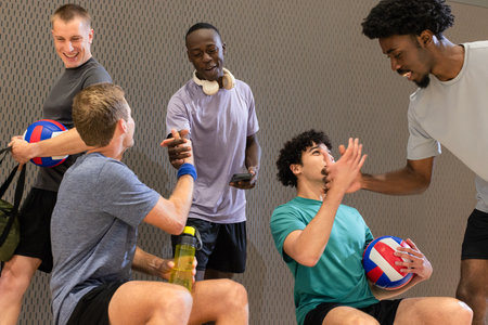 Diverse male volleyball teammates resting in sports hall holding volleyballs and water bottle....