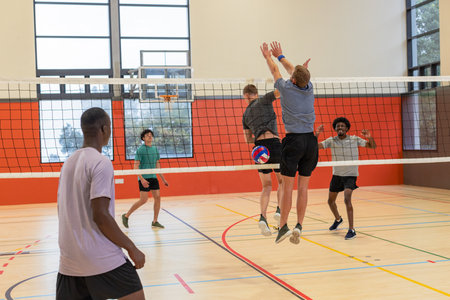Diverse male teammates jumping to block red white blue volleyball at gym net on wooden court. Action, sports, competition, teamwork, athleticism, vigor, recreationの写真素材