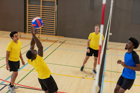 African American man jumping overhead pushing red-white-blue volleyball past net at gym with bench. Sports, teamwork, athleticism, energy, competition, fitness, activeの写真素材