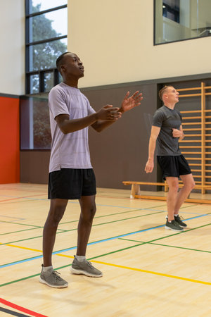 Diverse male teammates standing on gymnasium court near wooden wall bars practicing catching ball....