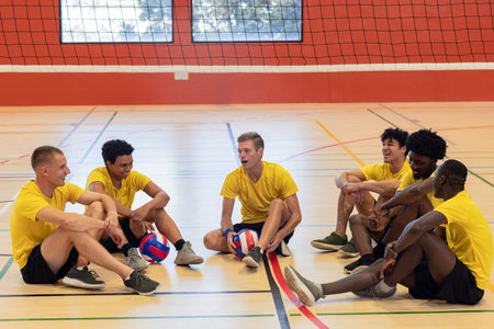 Diverse male volleyball team in uniforms sitting on court under net holding volleyballs. Athletes, camaraderie, teamwork, sportsmanship, active, energeticの写真素材