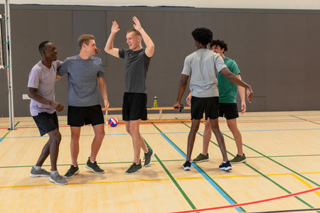 Diverse male teammates in athletic wear high-fiving while celebrating on gym court. Athletes, teamwork, activewear, recreation, fitness, competition, camaraderieの写真素材