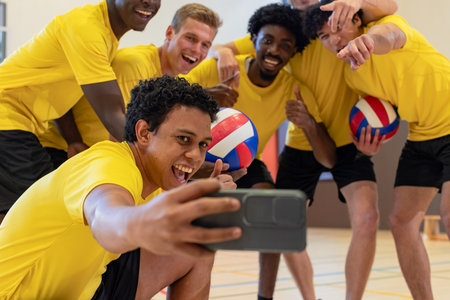 Diverse male volleyball teammates posing for group selfie on court with smartphone and volleyballs. Athletes, teamwork, camaraderie, competition, sport, fitness, recreationの写真素材