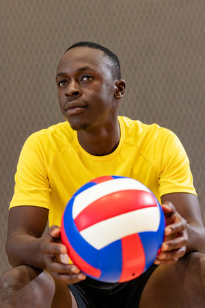 African American male athlete wearing sportswear holding volleyball and gazing upward at gym. Athlete, sports, fitness, contemporary, vibrant, determination, teamworkの写真素材