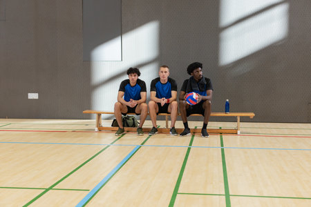 Diverse male teammates sitting in jerseys on bench in gym holding volleyball beside sports bag....
