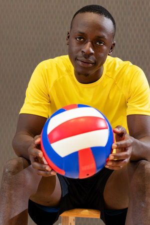 African American male athlete sitting on wooden bench in gym holding volleyball and gazing forward. Athlete, sports, competition, fitness, teamwork, training, dynamicsの写真素材