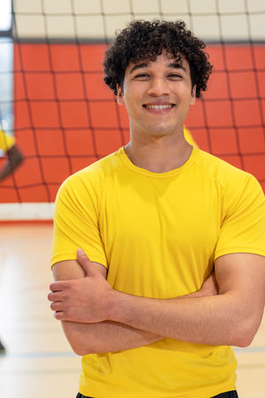 Male athlete standing with arms crossed before volleyball net while smiling inside gym court. Sport, gymnasium, athletic, teamwork, fitness, competition, vibrantの写真素材