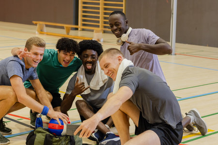 Diverse male teammates crouching on gym court checking smartphone next to volleyball and duffel bag. Athletes, teamwork, camaraderie, sporty, fitness, active, recreationalの写真素材