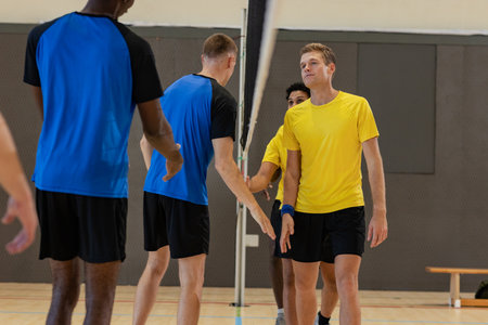 Diverse male volleyball players exchanging handshakes at net on gym court by sports bag, copy space. Athletic, teamwork, competition, sportsmanship, activewear, fitness, vibrantの写真素材