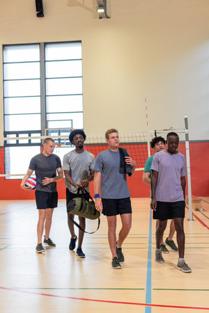 Diverse male teammates walking off court near volleyball net in gym carrying duffel bag and towel. Athletes, teamwork, active, camaraderie, recreational, fitness, modernの写真素材