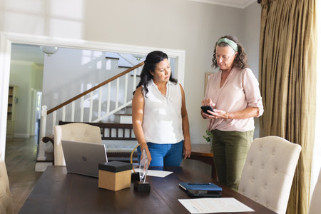 Two women discussing smartphone features at home office with laptop and documents. Technology,...