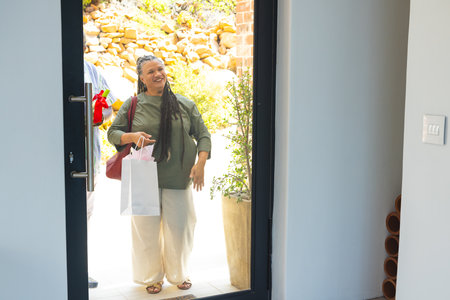 Smiling woman arriving with gift bag at home, ready for celebration. arrival, happiness, festive, cheerfulの写真素材