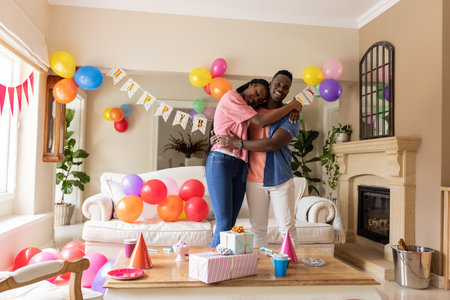Celebrating birthday together, African American couple hugging in decorated living room. Celebration, happiness, love, togetherness, decorations, joyfulの写真素材