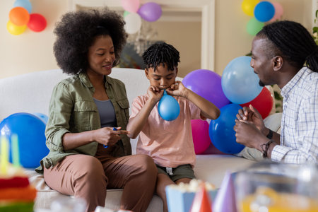 African American family celebrating at home, child blowing up balloon with parents. Celebration, togetherness, joy, cheerful, bonding, festiveの写真素材