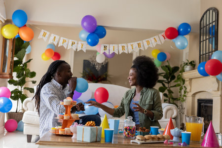 Celebrating birthday, African American couple with colorful balloons at party indoors. Celebration, festivity, joy, birthday cake, presents, happinessの写真素材