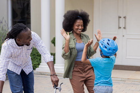 Child wearing helmet high-fiving mom, African American family playing outdoors. bonding, outdoor activity, parenting, childhood, recreation, togethernessの写真素材