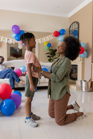 Mother kneeling, smiling at son in living room decorated with colorful balloons. Family,...