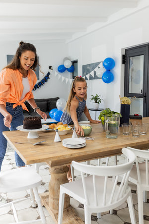 Mother and daughter setting table for birthday celebration at home, smiling happily. Family, party, festive, togetherness, parenthood, happinessの写真素材