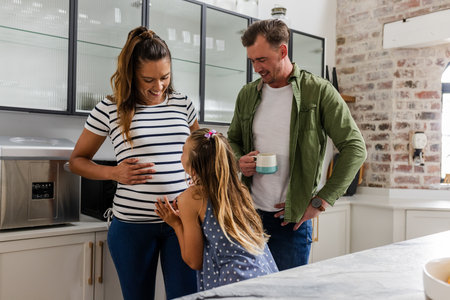 Expecting mother smiling as daughter kisses belly, father holding coffee mug nearby. Family, pregnancy, love, togetherness, parenting, bondingの写真素材