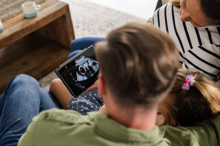 Family gathered on couch, viewing ultrasound image on tablet, sharing joyful moment. Pregnancy, expecting, technology, togetherness, love, parentingの写真素材