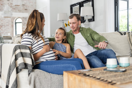Family relaxing on sofa, young girl touching pregnant mother's belly, smiling warmly. parenting, maternity, bond, love, living room, happinessの写真素材