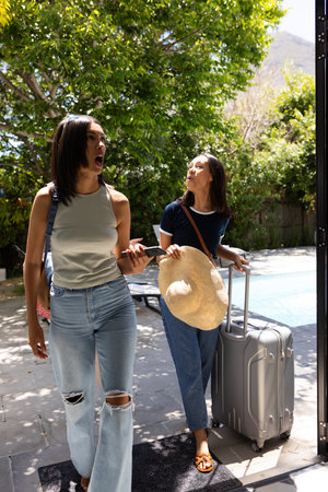 Arriving at sunny poolside, two young women appear excited and relaxed. They are carrying luggage and straw hat, ready for vacation getawayの写真素材