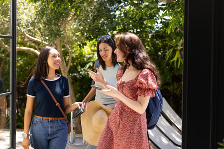 Chatting and smiling, three young women enjoy sunny day outdoors. They are discussing travel plans while holding suitcase and sun hatの写真素材