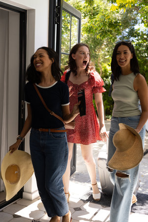 Three young women entering sunlit home, carrying hats and suitcase. They appear excited and ready for summer getaway, enjoying each other's companyの写真素材